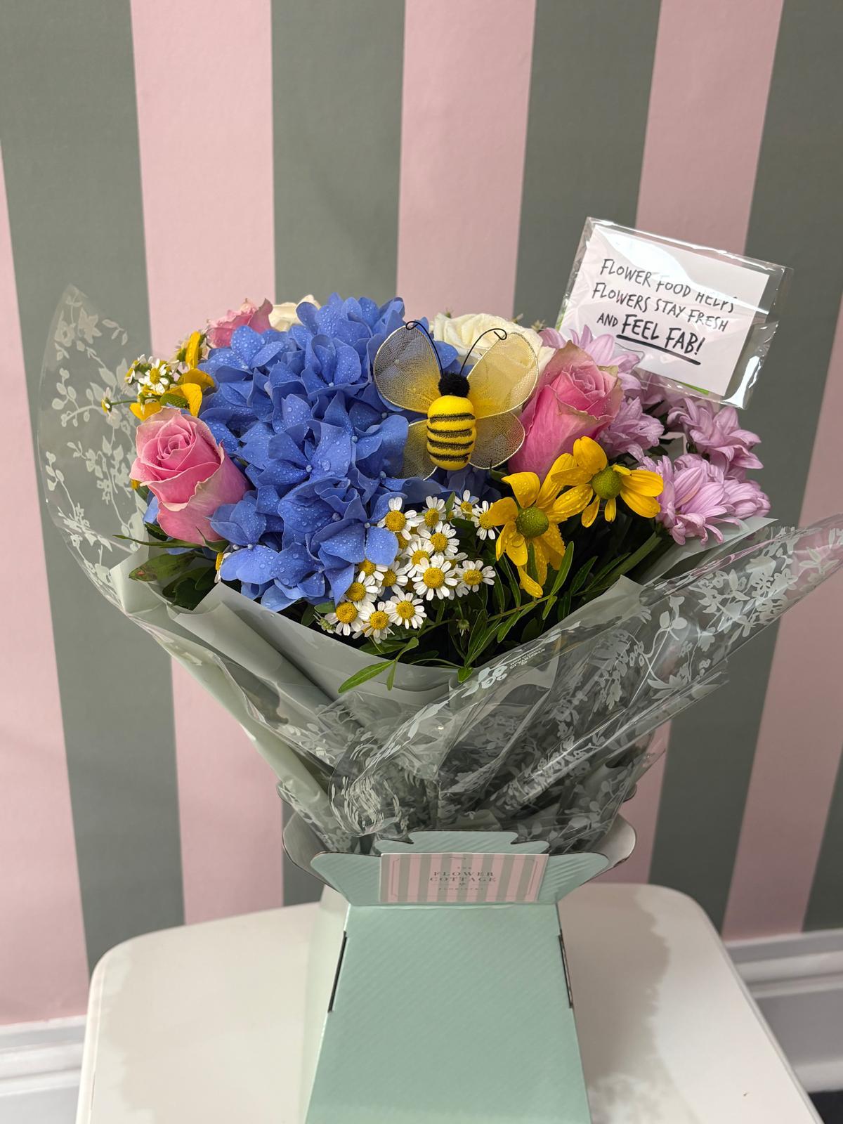 Blue hydrangeas, pink roses and yellow daisies with a decorative bee in a sage gift box